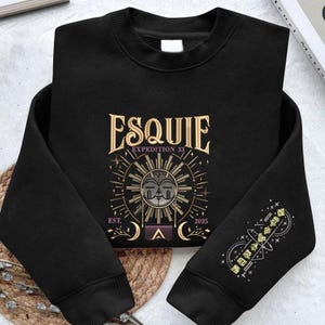 Puede incluir: Sudadera negra de cuello redondo con diseño gráfico dorado y morado. El diseño incluye la palabra "ESQUIE", "EXPEDITION 33", un sol y el año "2025". La manga presenta un diseño a juego. La sudadera está hecha de un material suave.