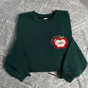 Peut inclure: Un sweat-shirt vert foncé avec un col rond et des manches longues. Le sweat-shirt présente une pomme brodée avec le texte "I PIPSQUEAK" au-dessus. La pomme a un visage souriant et est de style dessin animé.