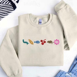 May include: A cream-colored sweatshirt with embroidered designs. The designs include a blue worm, a yellow wheat stalk, a blue fish, red cherries, a brown mouse, and a pink flower. The sweatshirt is made of a soft material.