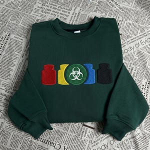 May include: Dark green sweatshirt with embroidered colorful graphics of jars and a biohazard symbol. The design includes red, yellow, blue, and black jars, and a green circle with a white biohazard symbol. The sweatshirt is folded on a newspaper background.
