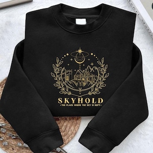 Puede incluir: Sudadera negra con un diseño gráfico dorado. El diseño presenta un castillo, montañas, una luna creciente, estrellas y la palabra "SKYHOLD". Debajo, el texto "THE PLACE WHERE THE SKY IS KEPT."