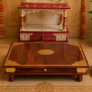 May include: A wooden prayer altar with a dark brown surface and gold accents. The altar has a drawer and is supported by four small, rounded legs. A smaller, white and red altar sits behind it, along with a brass lamp and a potted plant.