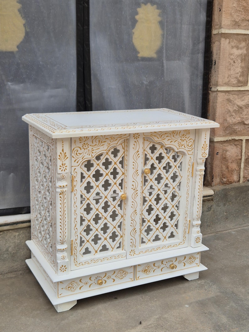 May include: A white cabinet featuring two doors with a lattice cutout pattern and gold painted floral accents. The piece includes two small drawers at the base with gold knobs and decorative gold painted details along the edges and frame.