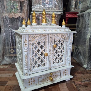 May include: A white wooden home temple featuring intricate gold painted floral patterns, lattice-style double doors with gold knobs, and a single drawer. The top is adorned with six gold-coloured finials arranged in a decorative architectural design.