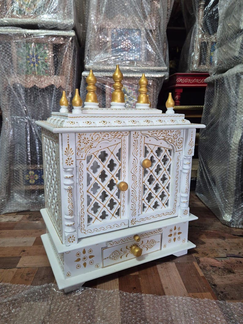 May include: A white wooden home temple featuring intricate gold painted floral patterns, lattice-style double doors with gold knobs, and a single drawer. The top is adorned with six gold-coloured finials arranged in a decorative architectural design.