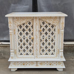May include: A white wooden cabinet featuring two doors with a gold-painted lattice pattern and two bottom drawers with gold floral accents. The cabinet has carved vertical pillars on each side and small round gold knobs on the doors and drawers.