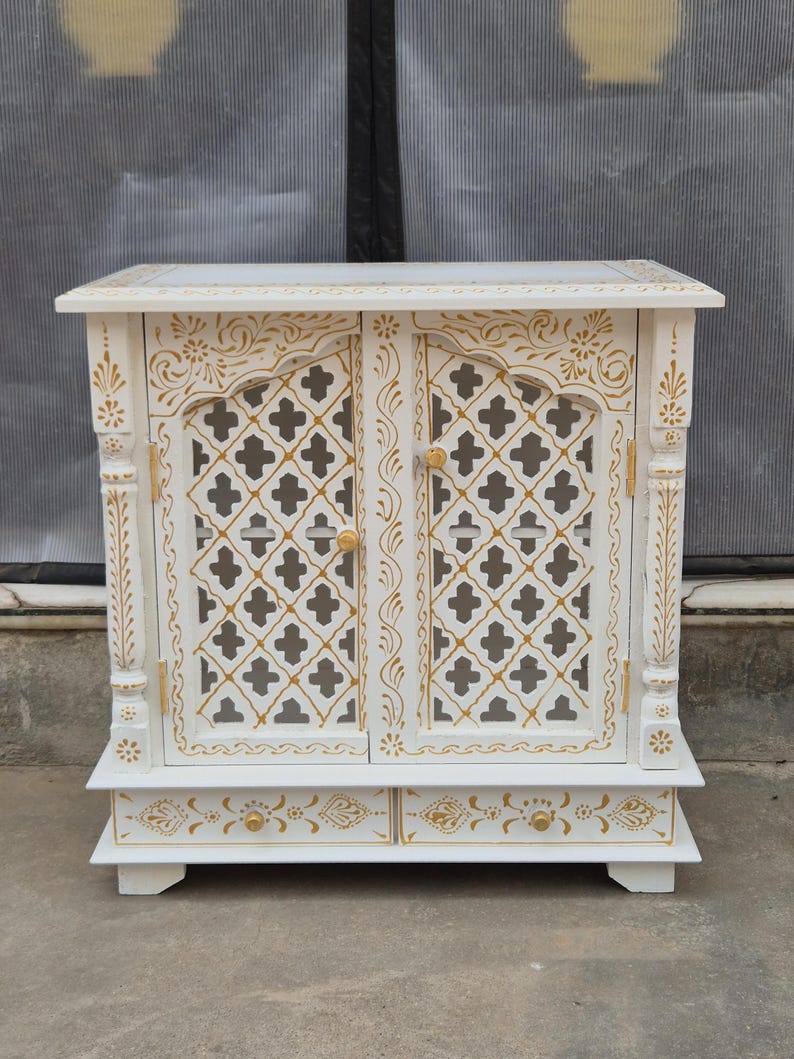 May include: A white wooden cabinet featuring two doors with a gold-painted lattice pattern and two bottom drawers with gold floral accents. The cabinet has carved vertical pillars on each side and small round gold knobs on the doors and drawers.