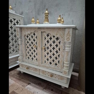 May include: A white wooden cabinet featuring intricate gold-painted floral patterns and lattice-style door panels. The structure includes two doors with small round knobs, a bottom drawer, and five decorative gold-toned finials on the top surface.