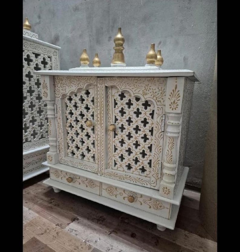 May include: A white wooden cabinet featuring intricate gold-painted floral patterns and lattice-style door panels. The structure includes two doors with small round knobs, a bottom drawer, and five decorative gold-toned finials on the top surface.