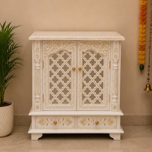 May include: A white wooden cabinet featuring two doors with intricate lattice cutouts and gold painted floral accents. The cabinet includes two lower drawers with gold knobs and decorative gold trim along the top and drawer fronts.