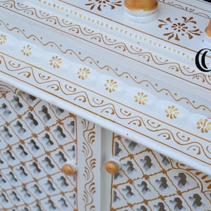 May include: A white wooden cabinet featuring intricate gold painted floral and scroll patterns. The cabinet has a lattice-style door design with small gold knobs and a scalloped edge along the top shelf. A black logo with the text CC is visible.