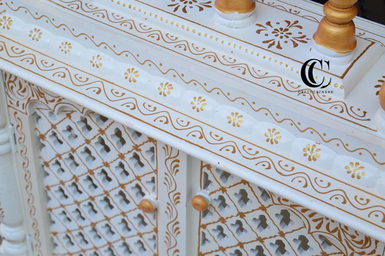 May include: A white wooden cabinet featuring intricate gold painted floral and scroll patterns. The cabinet has a lattice-style door design with small gold knobs and a scalloped edge along the top shelf. A black logo with the text CC is visible.