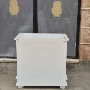 May include: A rectangular white wooden chest with a flat top surface featuring decorative light-colored trim along the edges. The front panel is solid white, and two silver-toned metal hinges are visible on the upper corners of the front face.