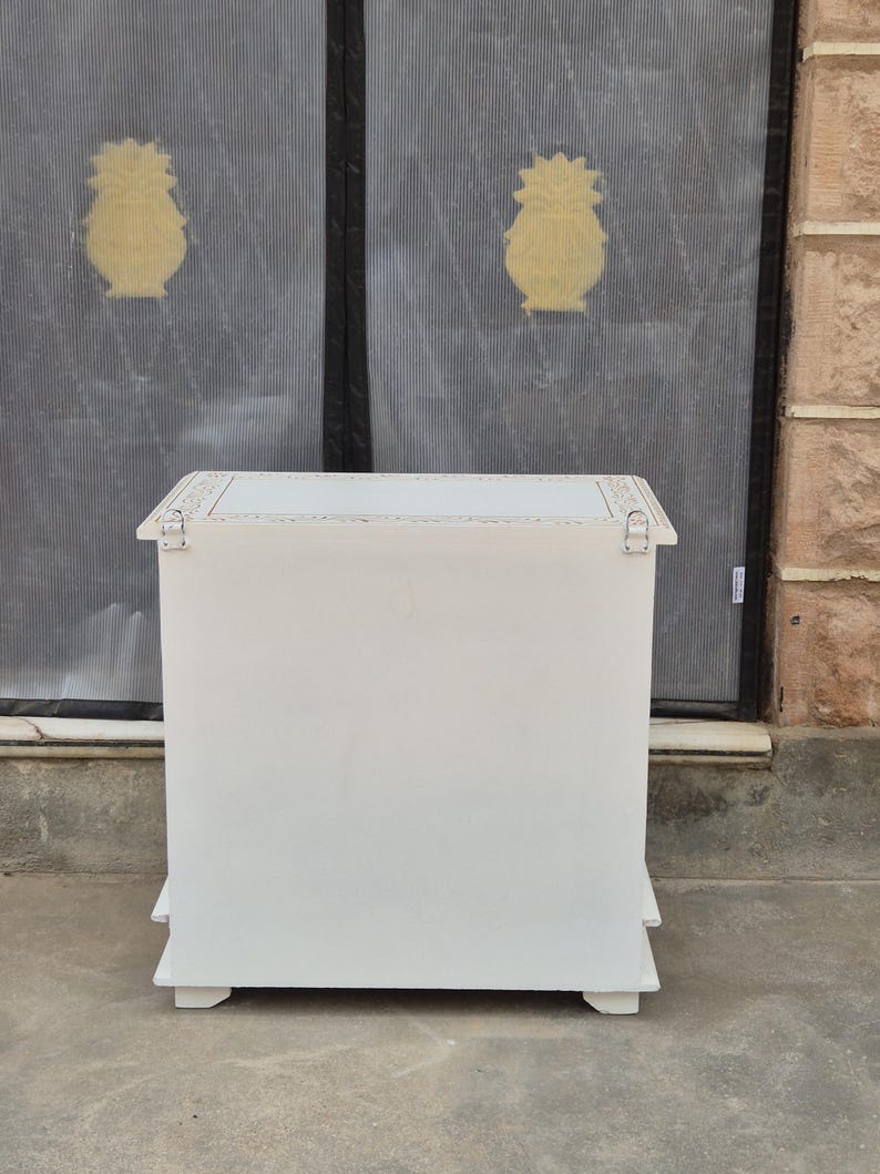 May include: A rectangular white wooden chest with a flat top surface featuring decorative light-colored trim along the edges. The front panel is solid white, and two silver-toned metal hinges are visible on the upper corners of the front face.