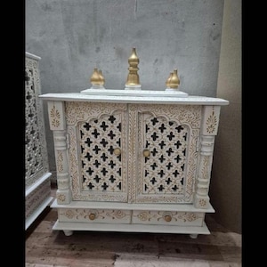 May include: A white wooden cabinet with two doors featuring intricate lattice cutouts and gold painted accents. The top surface holds three gold finials, and the base includes two small drawers with gold knobs. The structure has carved pillars.