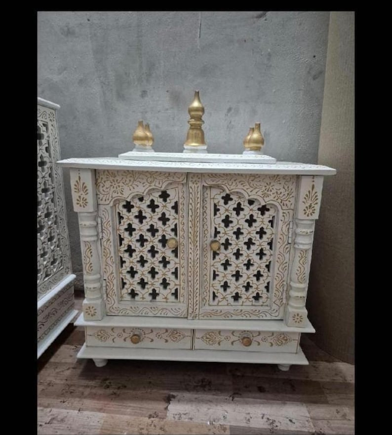 May include: A white wooden cabinet with two doors featuring intricate lattice cutouts and gold painted accents. The top surface holds three gold finials, and the base includes two small drawers with gold knobs. The structure has carved pillars.