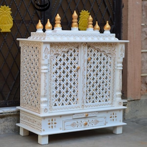 May include: A white wooden home temple featuring gold painted accents, intricate lattice door panels, and a pull-out drawer. The top is adorned with multiple gold finials, and the entire structure is decorated with detailed floral and geometric patterns.