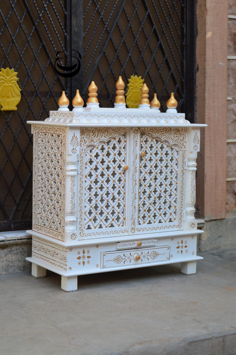 May include: A white wooden home temple featuring gold painted accents, intricate lattice door panels, and a pull-out drawer. The top is adorned with multiple gold finials, and the entire structure is decorated with detailed floral and geometric patterns.