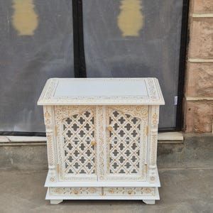 May include: A white wooden cabinet featuring two doors with gold-painted lattice patterns and two small drawers at the base. The cabinet has a flat top surface, carved vertical pillars on the sides, and small gold knobs on the doors and drawers.
