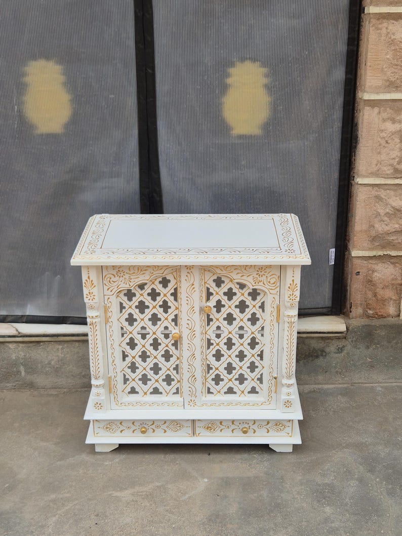 May include: A white wooden cabinet featuring two doors with gold-painted lattice patterns and two small drawers at the base. The cabinet has a flat top surface, carved vertical pillars on the sides, and small gold knobs on the doors and drawers.