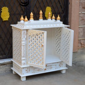 May include: A white wooden home temple cabinet featuring gold painted accents, intricate lattice work on the two open doors, and a small pull-out drawer at the base. The top is adorned with six gold finials and decorative carved patterns.