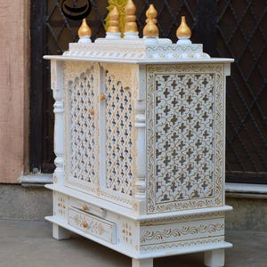May include: A white wooden home temple featuring intricate gold-painted floral patterns and lattice-style doors. The structure includes a pull-out drawer with a small knob, four short legs, and a top adorned with five gold-tipped decorative finials.