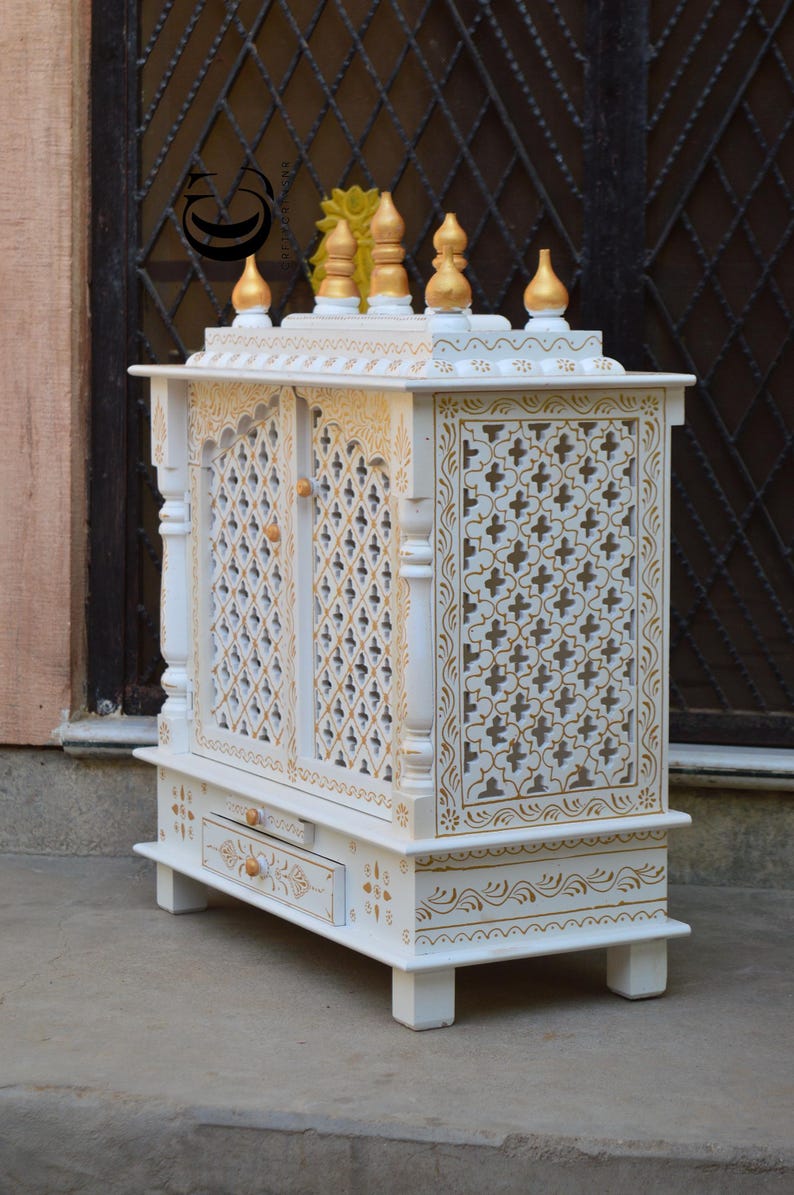 May include: A white wooden home temple featuring intricate gold-painted floral patterns and lattice-style doors. The structure includes a pull-out drawer with a small knob, four short legs, and a top adorned with five gold-tipped decorative finials.