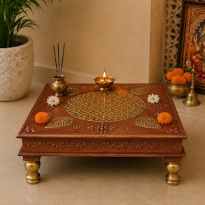 May include: A square, wooden altar table with a detailed, carved design. The table has gold-coloured accents, including the legs and a central decorative pattern. A small oil lamp is lit on top, along with incense and flowers.