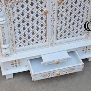 May include: A white wooden cabinet featuring gold painted floral and geometric patterns. The cabinet has two doors with a lattice design, small gold knobs, and a pull-out drawer at the base. A logo with the letters CC is visible on the right door.