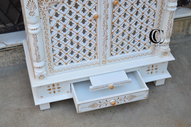 May include: A white wooden cabinet featuring gold painted floral and geometric patterns. The cabinet has two doors with a lattice design, small gold knobs, and a pull-out drawer at the base. A logo with the letters CC is visible on the right door.