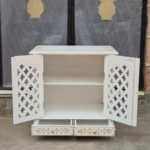 May include: A white wooden cabinet featuring two open doors with intricate cross-shaped cutouts and gold painted accents. The cabinet includes two lower drawers with gold floral patterns and small round gold knobs, providing storage space.