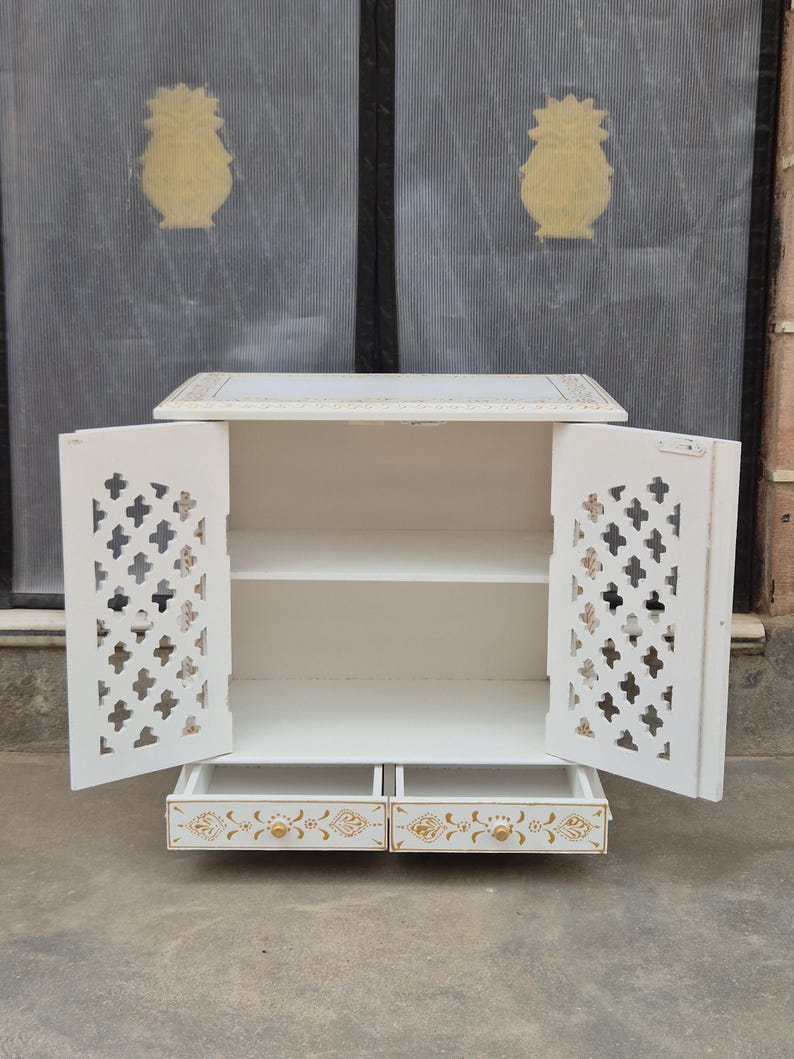 May include: A white wooden cabinet featuring two open doors with intricate cross-shaped cutouts and gold painted accents. The cabinet includes two lower drawers with gold floral patterns and small round gold knobs, providing storage space.