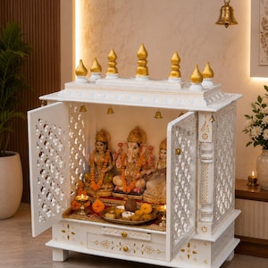 May include: A white wooden home temple featuring intricate lattice doors, a drawer, and a tiered roof with gold-painted finials. Inside, three deity statues are arranged on a red cloth, accompanied by small brass oil lamps and a metal offering tray.