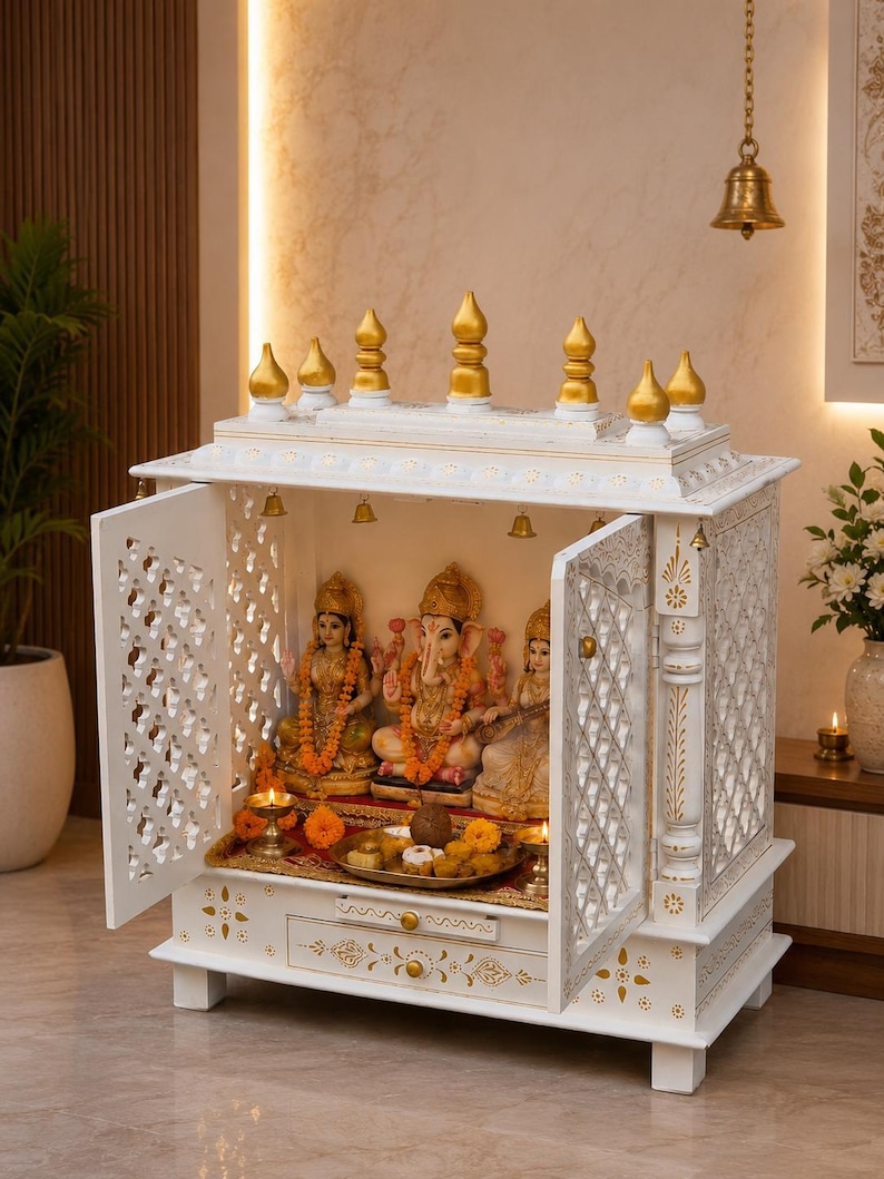 May include: A white wooden home temple featuring intricate lattice doors, a drawer, and a tiered roof with gold-painted finials. Inside, three deity statues are arranged on a red cloth, accompanied by small brass oil lamps and a metal offering tray.