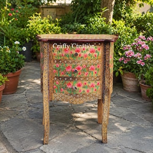 May include: A wooden bedside table with three drawers, featuring hand-painted pink floral motifs and intricate dark brown scrollwork patterns on the frame and legs. The text Crafty Creation is visible on the top drawer front.