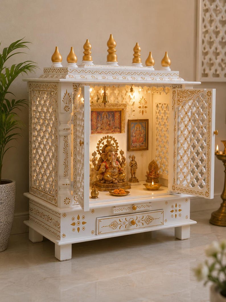 May include: A white wooden home temple featuring gold-painted accents, intricate lattice side panels, and a pull-out drawer. The interior houses a central Ganesha statue, smaller deity figures, framed religious prints, and a small hanging brass bell.