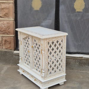 May include: A white wooden cabinet featuring two doors with intricate lattice cutouts and gold painted detailing. The cabinet includes a small drawer at the base with a gold knob, and the doors are also fitted with small gold knobs for opening.