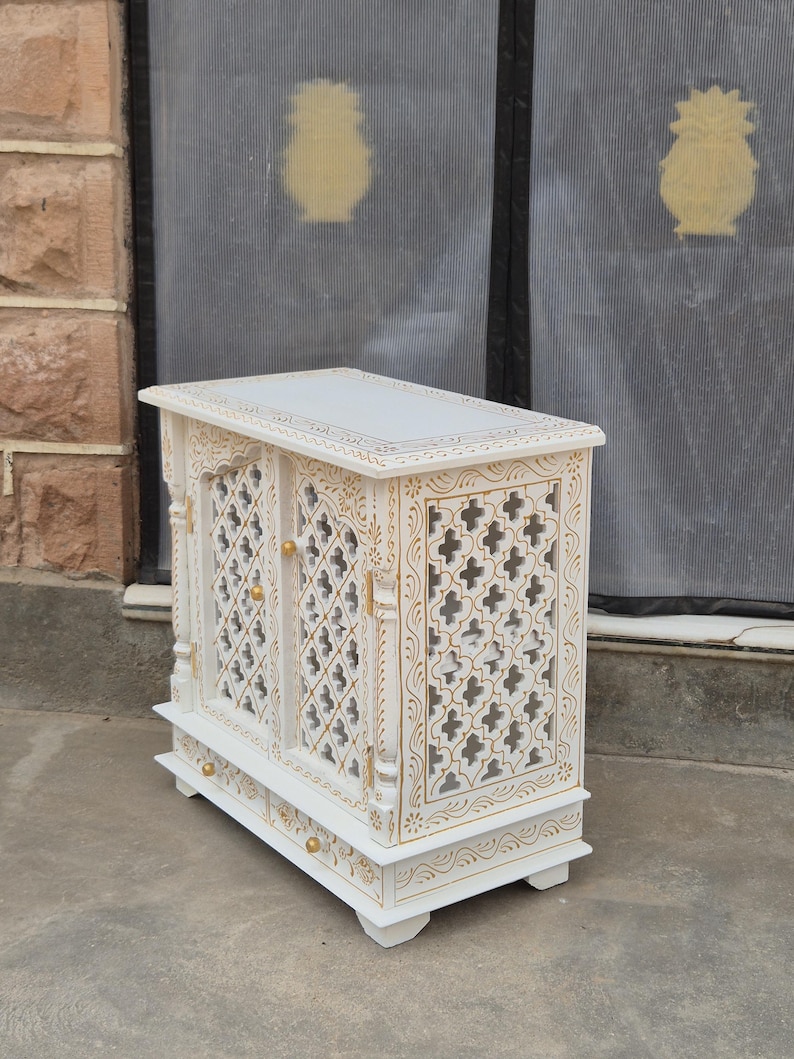 May include: A white wooden cabinet featuring two doors with intricate lattice cutouts and gold painted detailing. The cabinet includes a small drawer at the base with a gold knob, and the doors are also fitted with small gold knobs for opening.