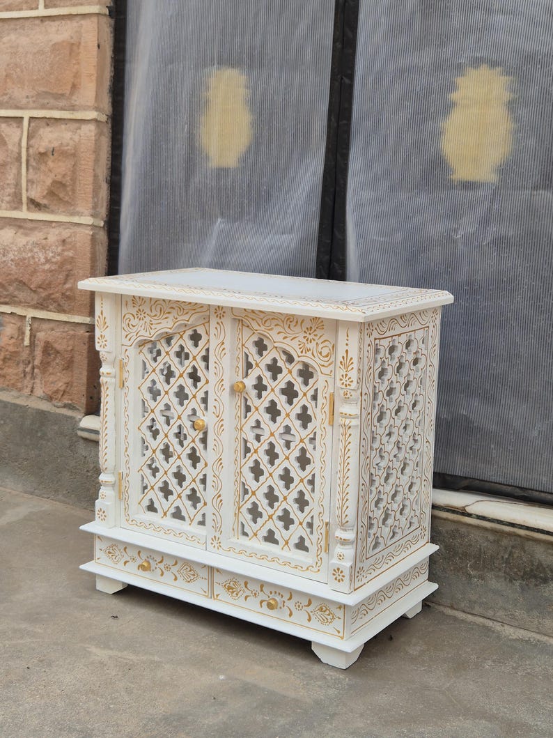 May include: A white wooden cabinet featuring two doors with a lattice cutout pattern and gold painted floral accents. The cabinet includes a single drawer at the base with two gold knobs and decorative gold detailing along the edges and frame.