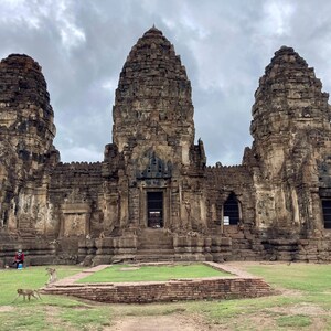 Puede incluir: Un gran complejo de templos antiguos con tres altas torres de piedra desgastada bajo un cielo nublado. La estructura presenta múltiples niveles, puertas y ventanas. Monos en la hierba verde frente al templo.