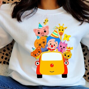 May include: White sweatshirt with a vibrant cartoon design of characters in a yellow car. The graphic includes a pig, bear, giraffe, bee, and other animals wearing party hats. The car has a red nose and black wheels.