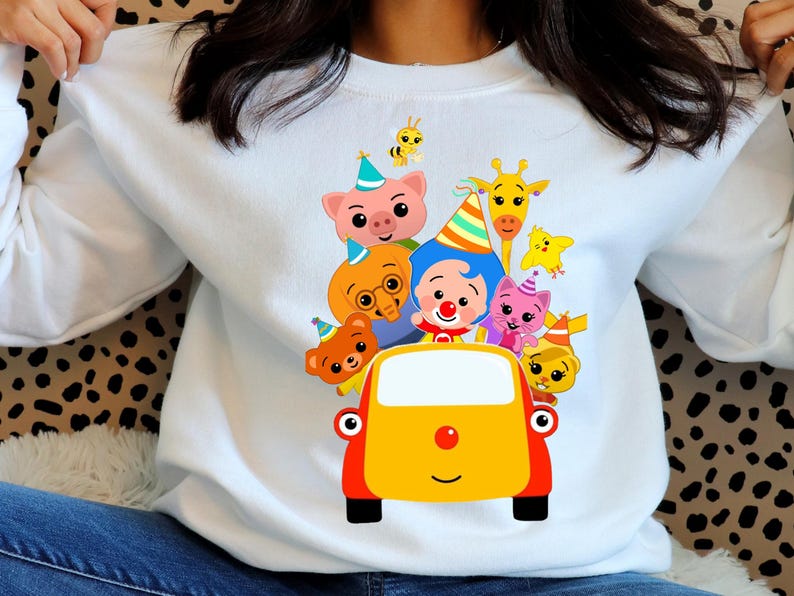 May include: White sweatshirt with a vibrant cartoon design of characters in a yellow car. The graphic includes a pig, bear, giraffe, bee, and other animals wearing party hats. The car has a red nose and black wheels.