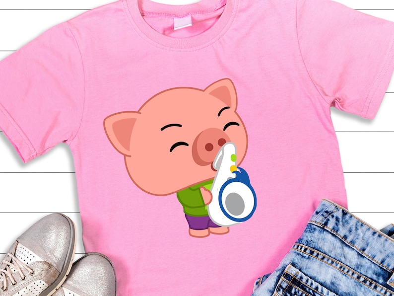 May include: Pink t-shirt featuring a cartoon pig wearing a green shirt and purple pants, holding a white and blue object. The pig has a smiling expression. The shirt is displayed with silver shoes and blue jeans.