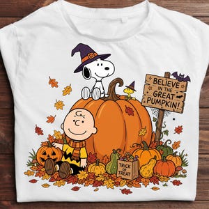 May include: A white t-shirt featuring a graphic of Snoopy in a witch hat and Charlie Brown sitting among orange pumpkins and autumn leaves. A wooden sign reads BELIEVE IN THE GREAT PUMPKIN! and a small bag is labeled TRICK OR TREAT.