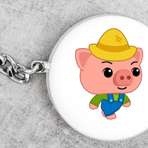 May include: A round keychain with a cartoon pig wearing a yellow hat, green shirt, and blue overalls. The pig has a pink face and a smiling expression. The keychain has a silver chain and ring.
