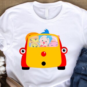 May include: White t-shirt featuring a colorful cartoon car with passengers. The car is yellow and red, with black wheels and large red eyes. Inside, there are several cartoon characters with different expressions.