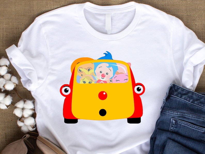 May include: White t-shirt featuring a colorful cartoon car with passengers. The car is yellow and red, with black wheels and large red eyes. Inside, there are several cartoon characters with different expressions.