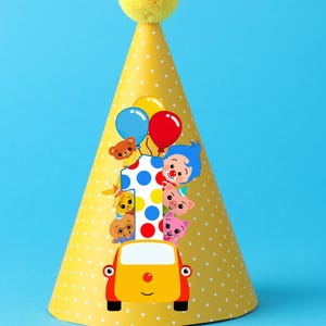 Op de afbeelding: Een gele feesthoed met witte stippen en een gele pompon. De hoed heeft een kleurrijke afbeelding van een auto met stripfiguren, ballonnen en het nummer 1. De achtergrond is effen blauw.