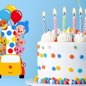 Op de afbeelding: Een witte verjaardagstaart versierd met kleurrijke stippen, witte glazuurkrullen en brandende kaarsen. Aan de linkerkant een kleurrijke &eacute;&eacute;n met stripfiguren en ballonnen.