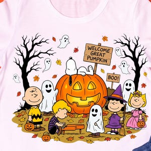 May include: A white t-shirt featuring a Peanuts Halloween graphic with Snoopy on a large orange jack-o'-lantern, Charlie Brown, Linus, Lucy in a witch hat, and Sally. The scene includes ghosts, bare trees, fallen leaves, and signs reading WELCOME GREAT PUMPKIN and BOO.